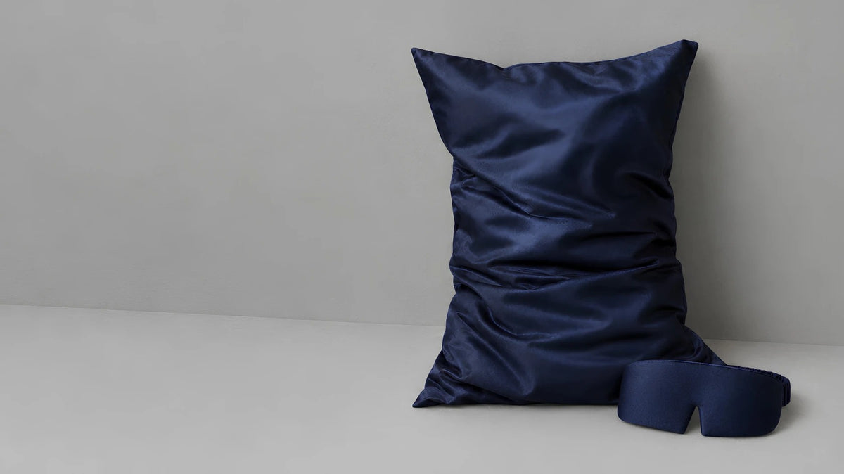navy blue silk pillowcase and silk sleep mask against a grey wall