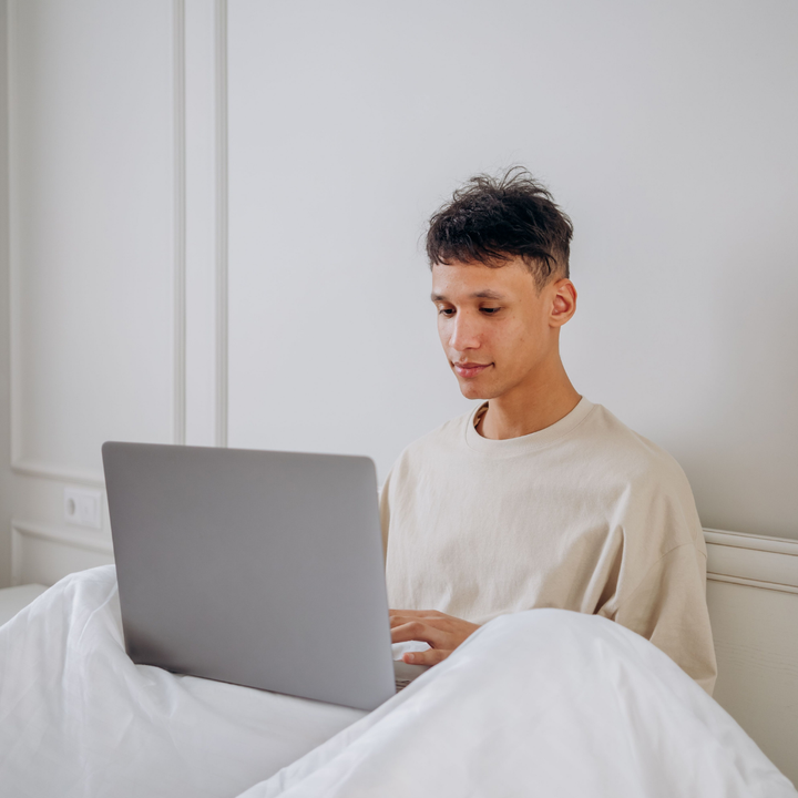 Junger Mann sitzt im Bett unter weißer Decke und recherchiert am Laptop, welcher Nasenstrip der beste ist