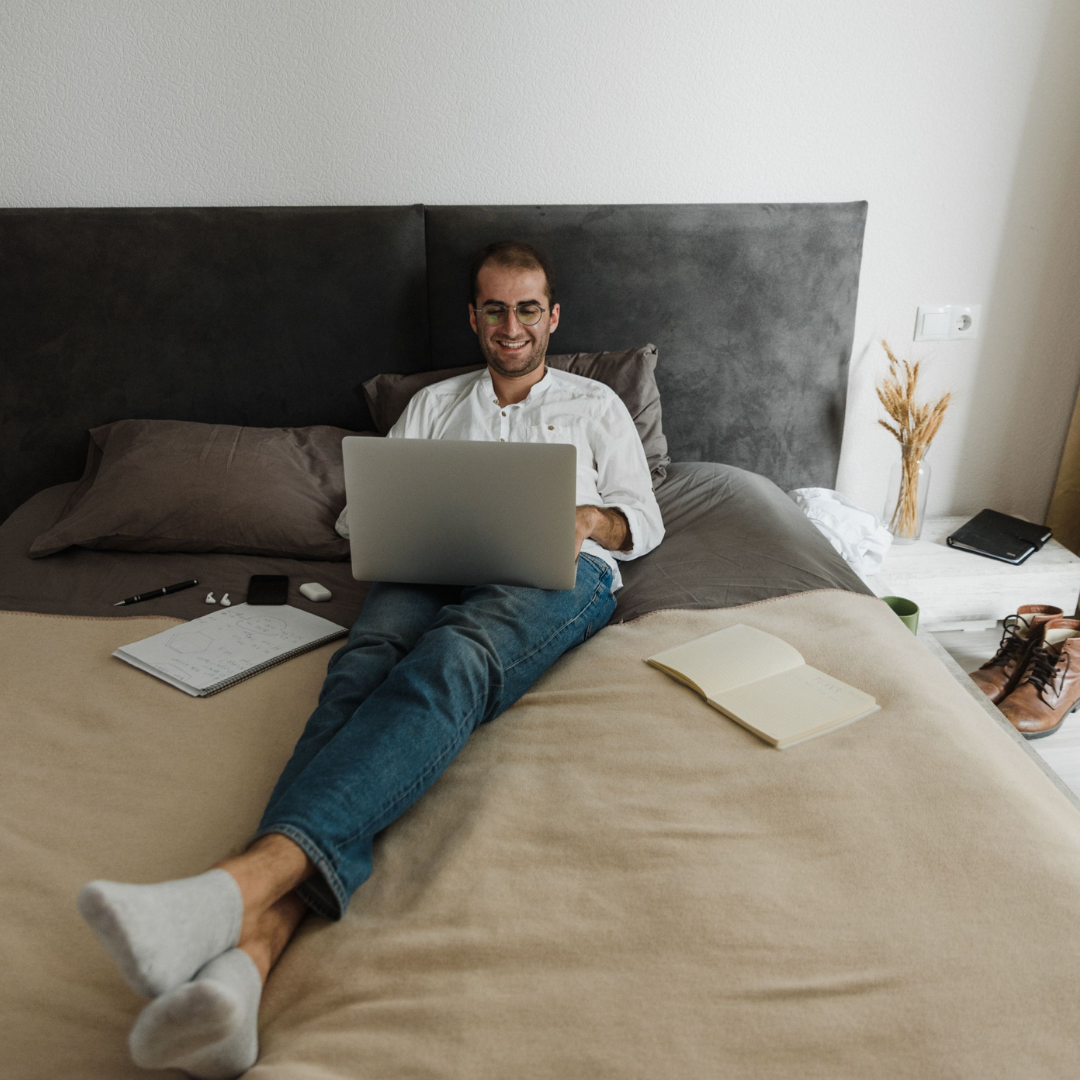Lächelnder Mann mit Brille sitzt mit Laptop auf dem Bett, Thema Mouth Taping oder Nasenstrips
