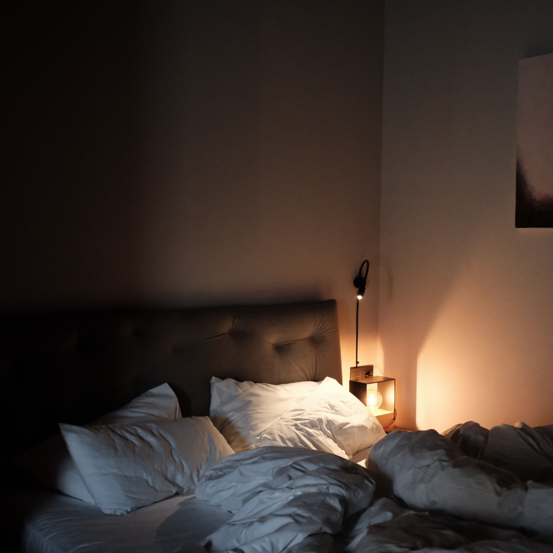 Dunkles Schlafzimmer mit zerwühltem weißem Bett und warmer Leselampe, Thema Atemstrips für die Nacht statt Porenstrips