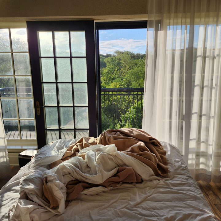 Zerwühltes Bett vor einer offenen Balkontür mit Morgenlicht und grünen Bäumen, Sinnbild für erholsamen Schlaf