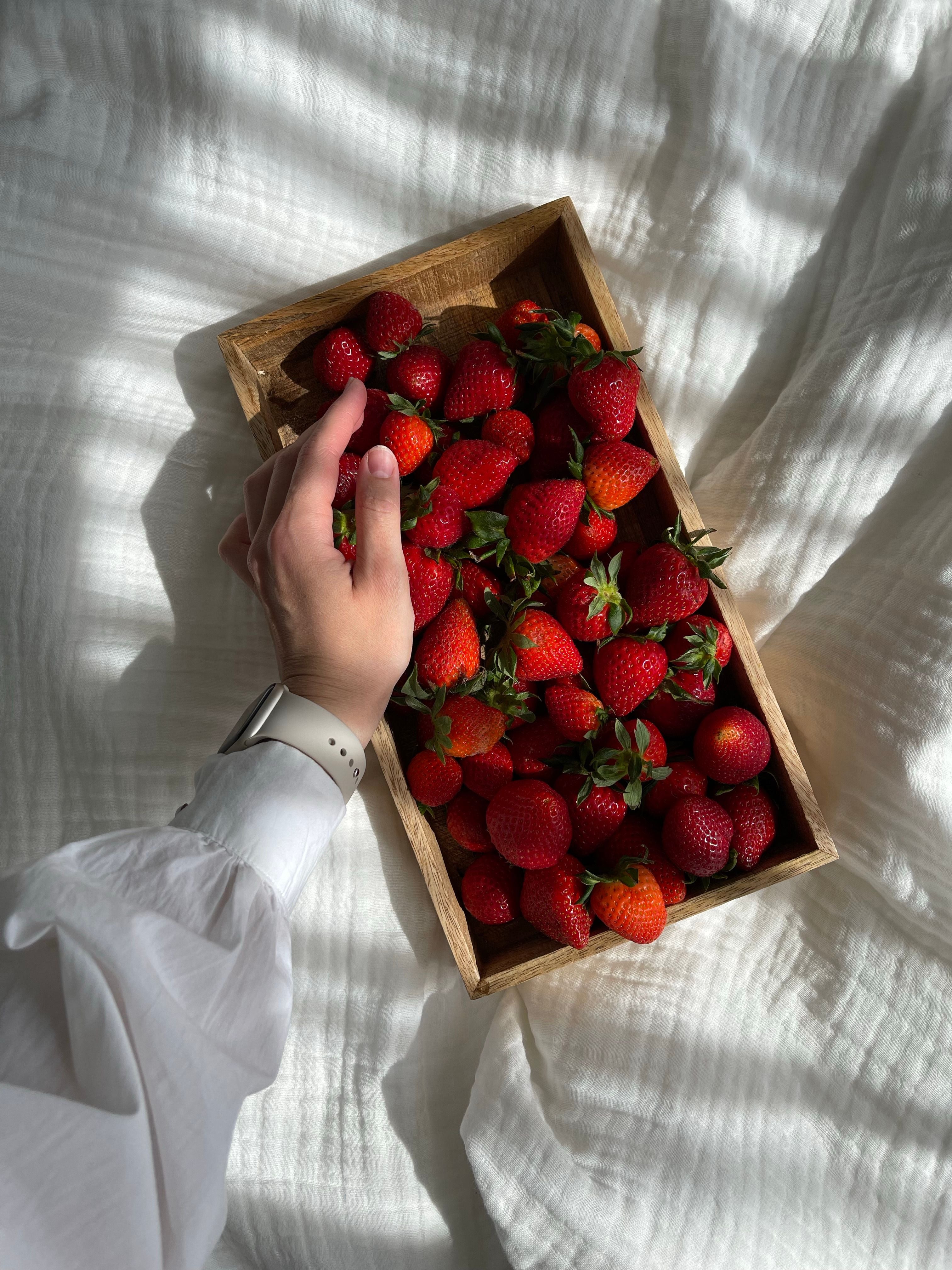 Hand greift nach frischen Erdbeeren in einer Holzkiste auf weißer Bettwäsche, Ernährung als Säule der Longevity