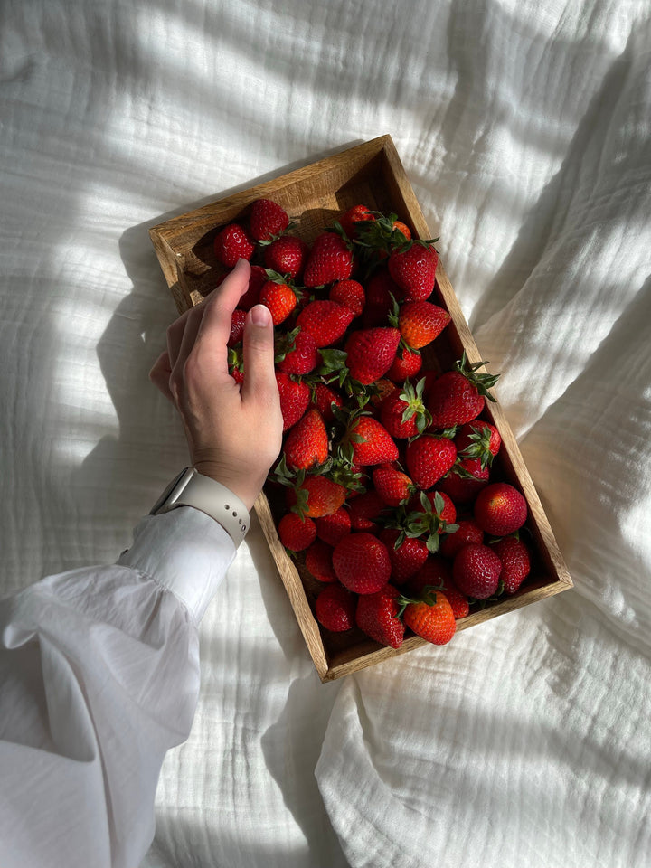 Hand greift nach frischen Erdbeeren in einer Holzkiste auf weißer Bettwäsche, Ernährung als Säule der Longevity