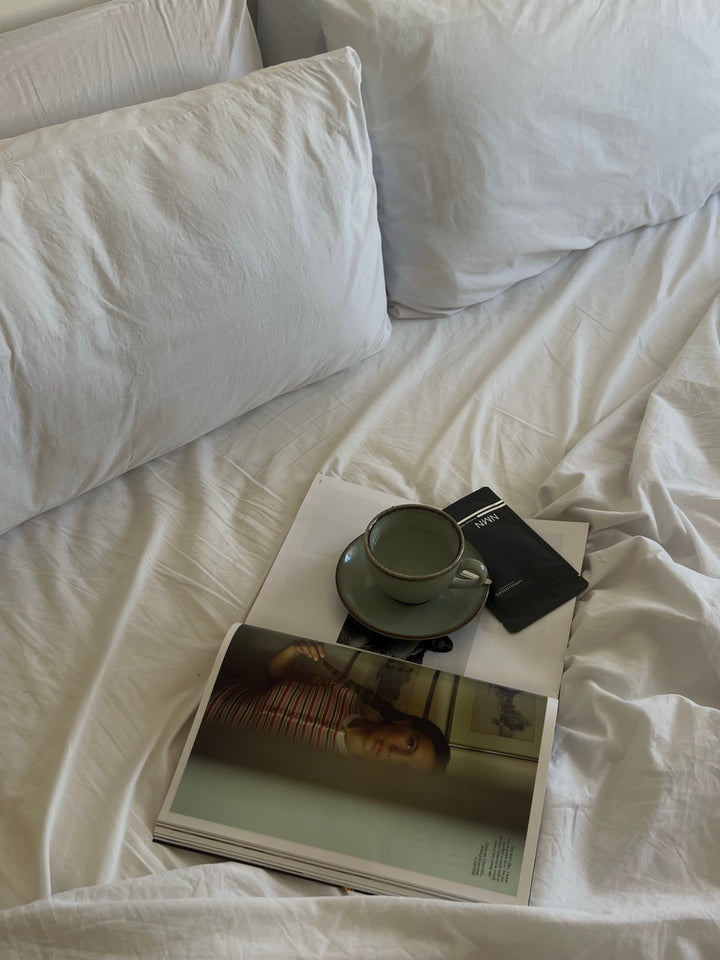 NMN Beutel, Tasse und aufgeschlagenes Magazin auf weißer Bettwäsche am Morgen