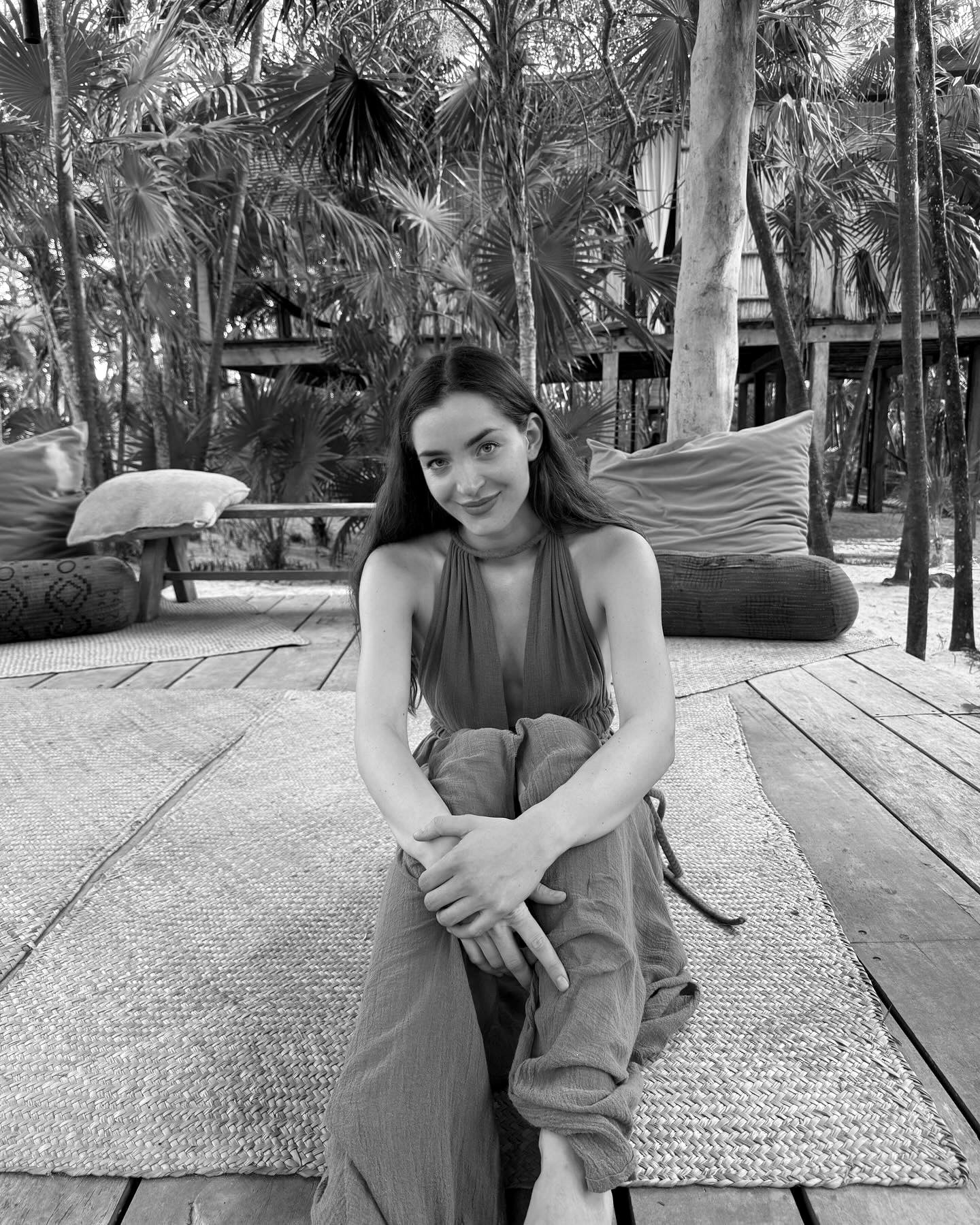 A woman is seated on a wooden deck, facing a tree that adds a natural element to the serene setting.