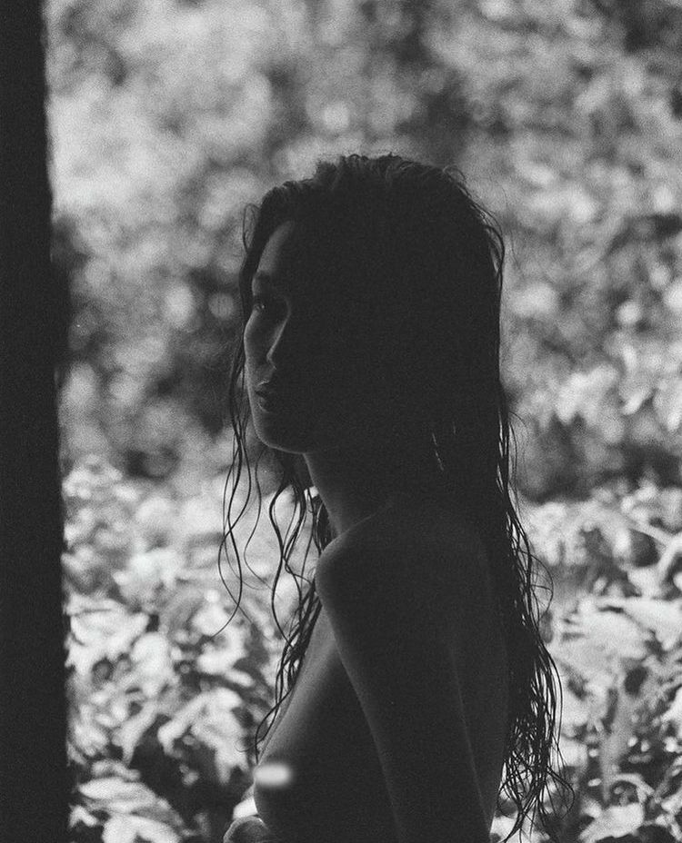 A black and white image of a person with long, wet hair standing outdoors, surrounded by blurred foliage. They are turned slightly to the side, with their silhouette and facial features partially in shadow.