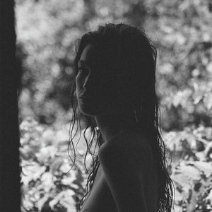 A black and white image of a person with long, wet hair standing outdoors, surrounded by blurred foliage. They are turned slightly to the side, with their silhouette and facial features partially in shadow.