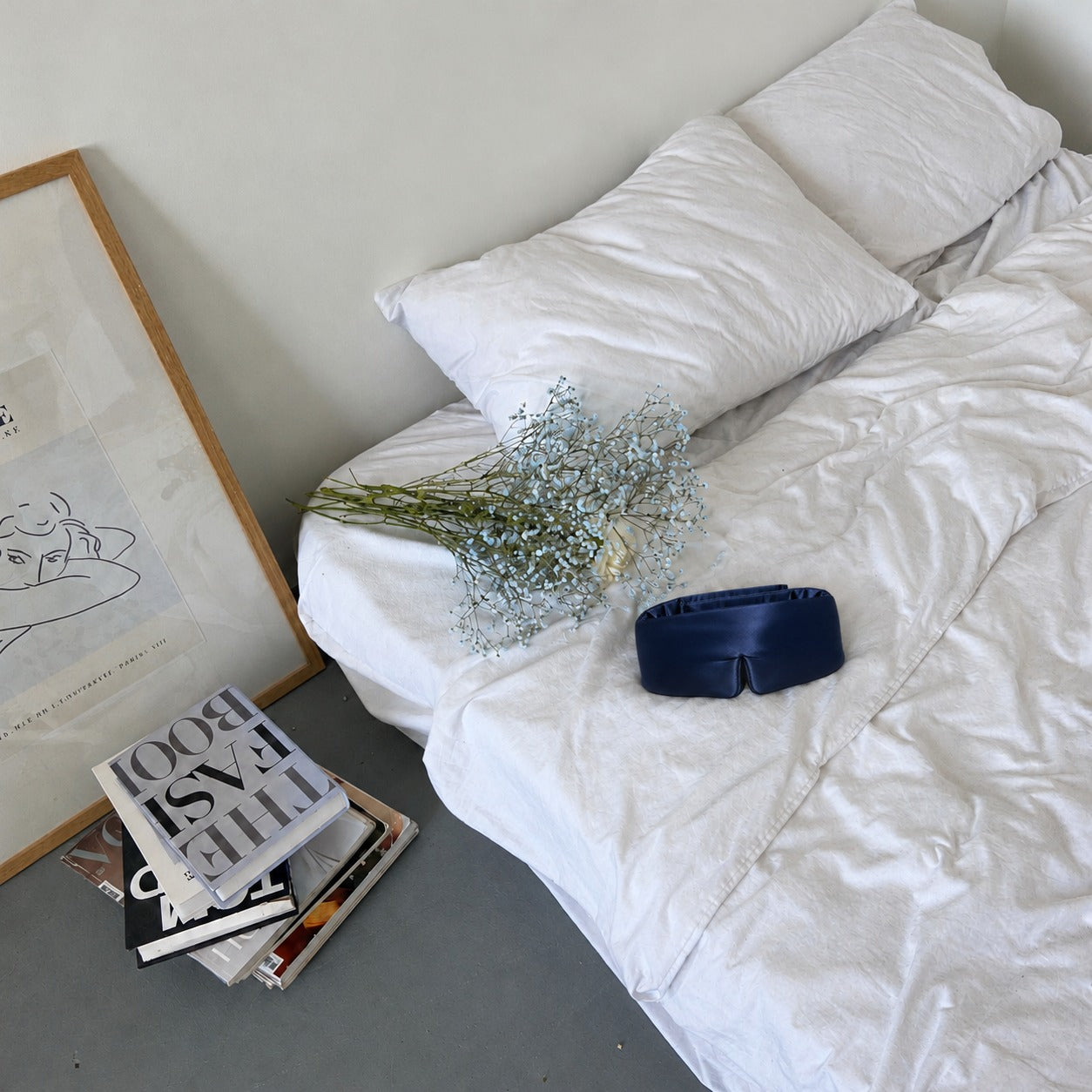 Silk-Schlafmaske in Dunkelblau auf einem Bett mit weißer Bettwäsche und Schleierkraut